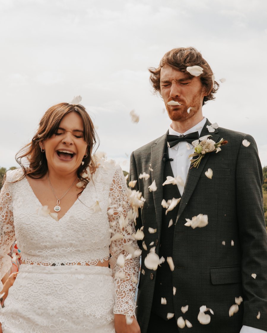 A laughing bride and groom showered in white petal confetti as guests cheer