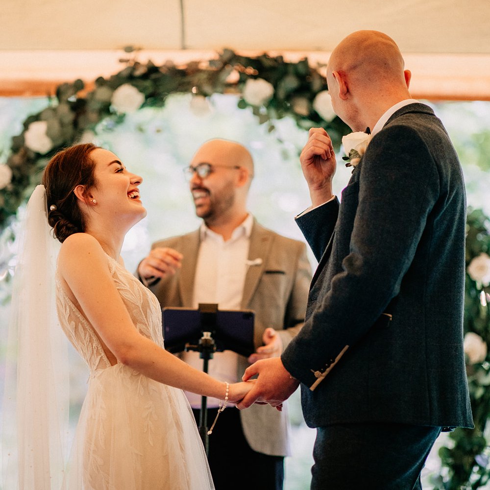 A bride and groom laughing as they hold hands under a flower arch, with Chris leading the ceremony